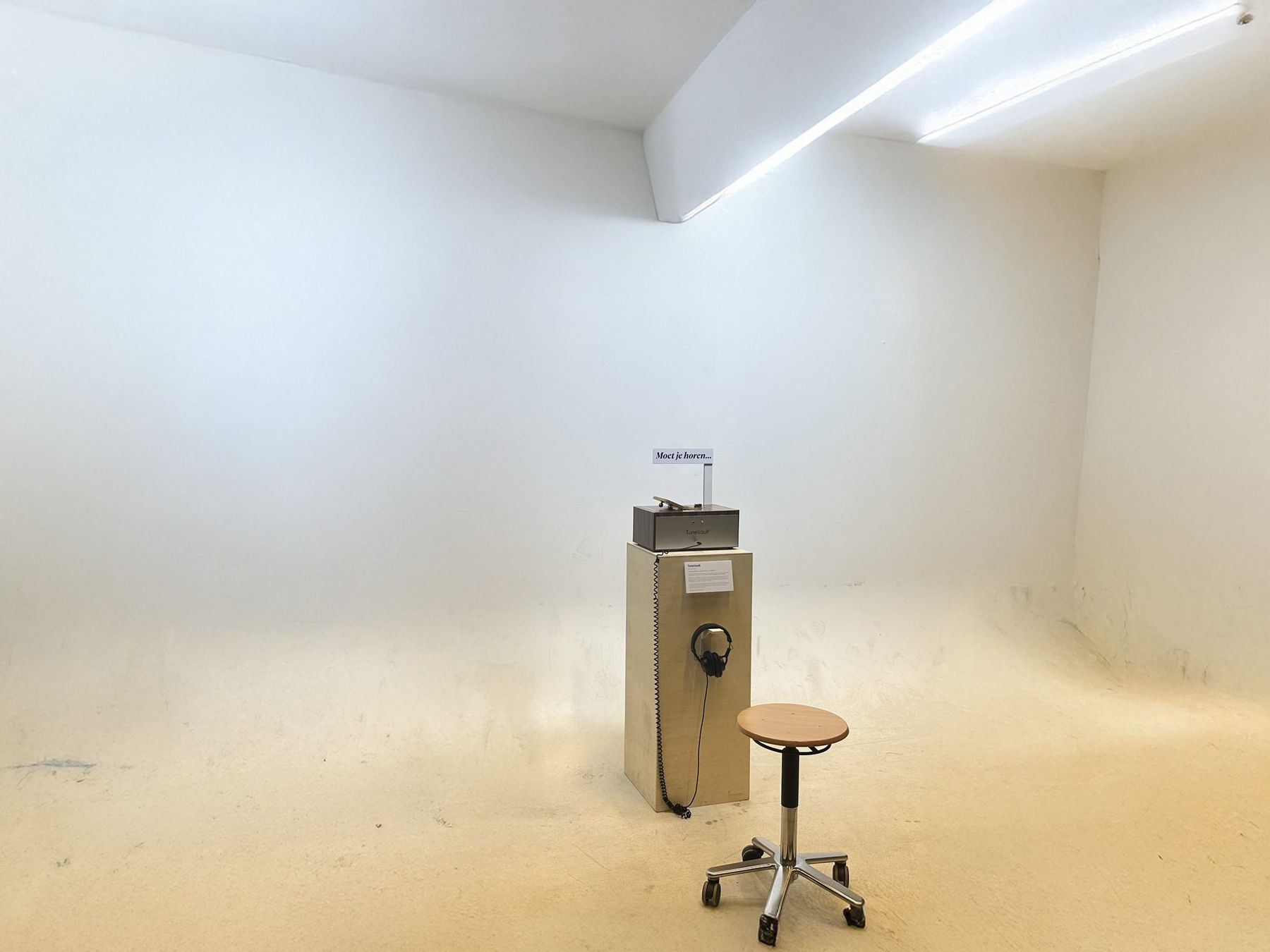 TuneVault on a plinth in an empty white exhibition room, with headphones hanging on the plinth and a stool in front