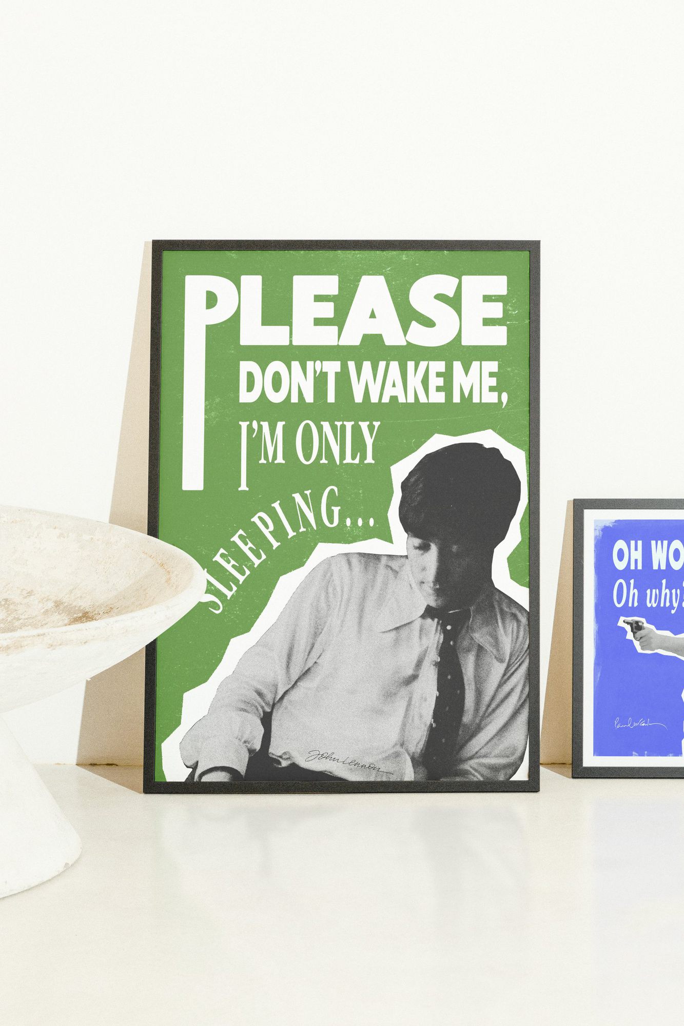 The John Lennon poster in a black frame, leaning on a white surface next to a bowl